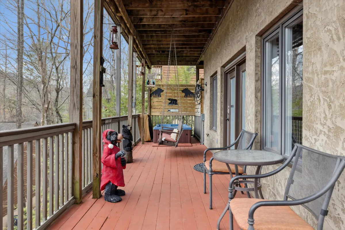 A welcoming porch area with a wooden railing, outdoor seating, and hanging lanterns, accented by a distinctive red bear statue. The space invites relaxation and casual conversation, surrounded by leafy trees.
