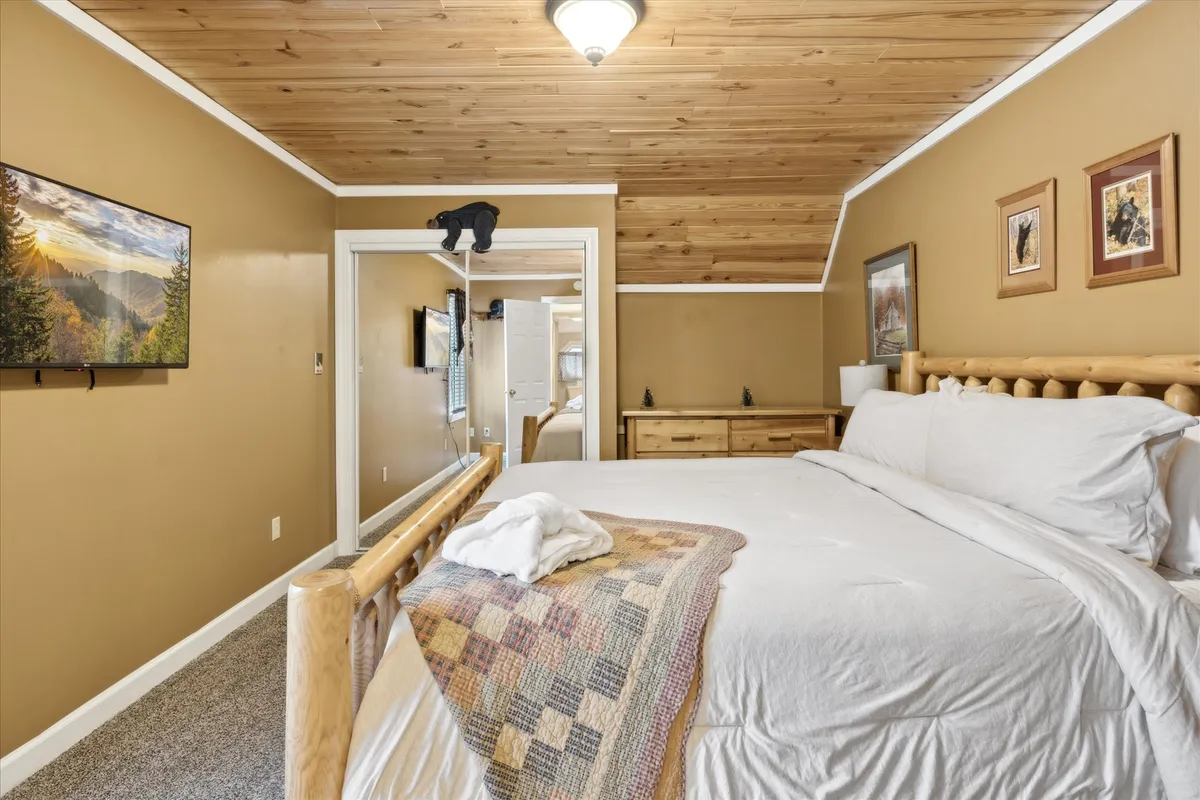 A warm, rustic bedroom featuring a wooden bed frame topped with a plaid throw blanket. The room includes a wall-mounted TV, a large mirror that creates an open feel, a wooden dresser, and framed art pieces. The soft lighting creates an inviting and restful atmosphere, perfect for unwinding after a day in the mountains.