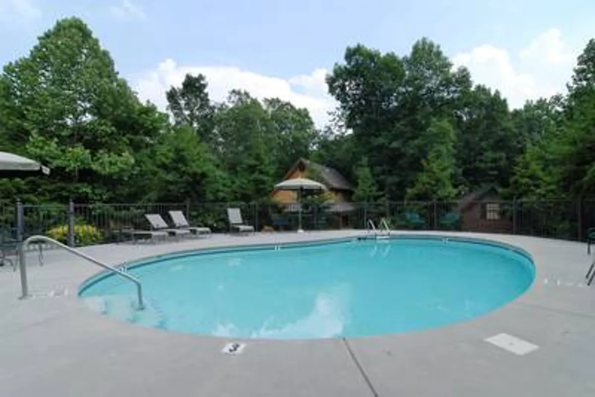 The image showcases the inviting exterior of a Pigeon Forge cabin, with a sparkling swimming pool surrounded by lounge chairs, an umbrella for shade, and lush trees creating a private, tranquil setting. The scene suggests relaxation at any time of day, with space for swimming, sunbathing, and social gatherings in nature.