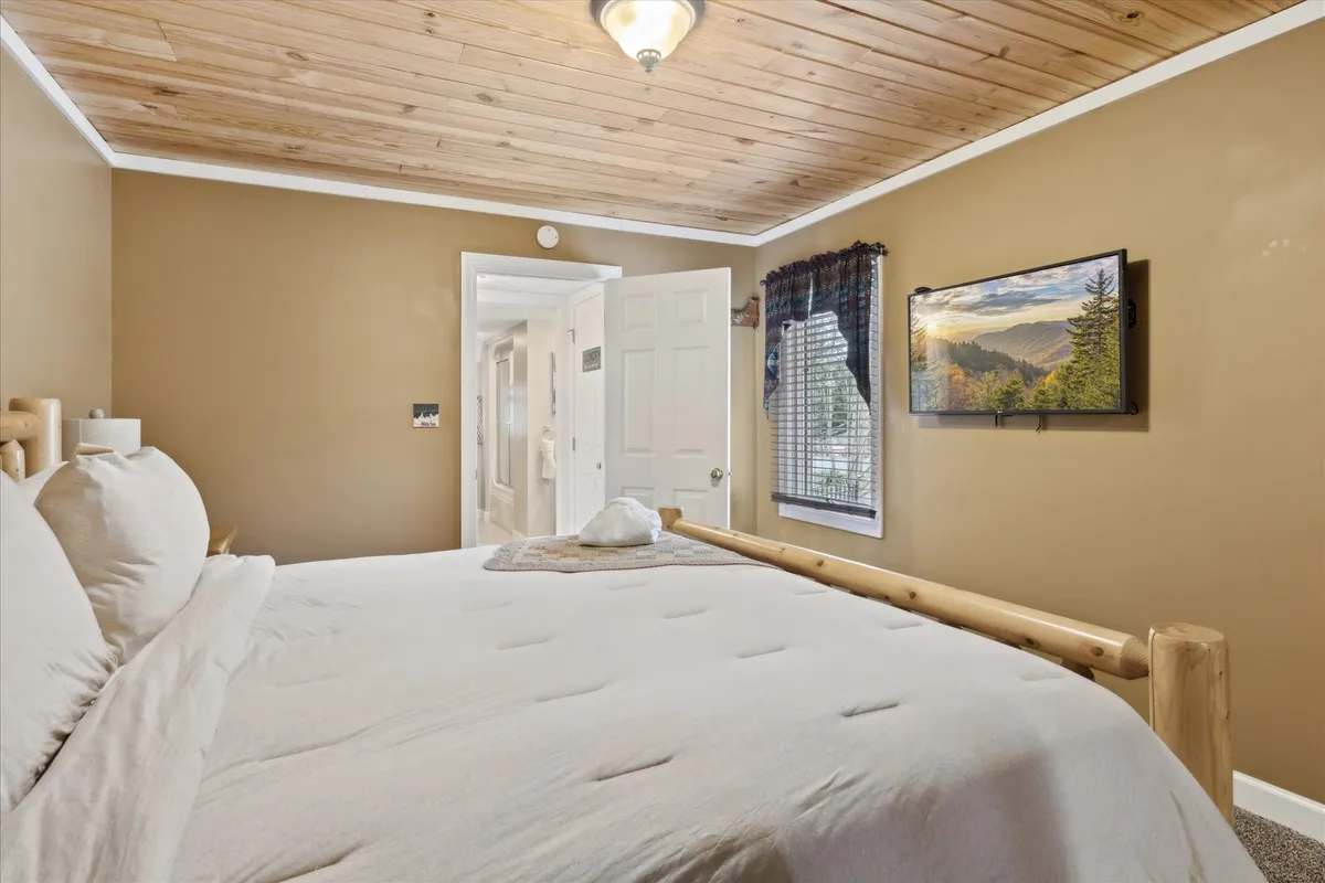 A serene bedroom with a bed dressed in crisp white linens set against a wooden frame. A large window with curtains lets in natural light, while a framed landscape picture adds a tranquil touch. The overall mood is calm and cozy, emphasizing peace and privacy.