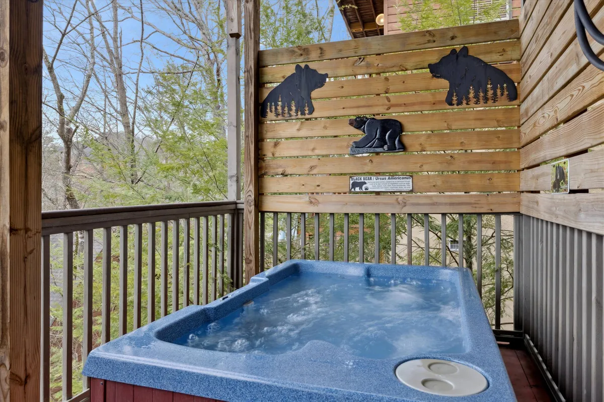 A tranquil outdoor hot tub area surrounded by a wooden fence with playful bear cutouts, nestled amidst lush trees. People are seen relaxing in the hot tub, enjoying the view of nature and sharing moments together.