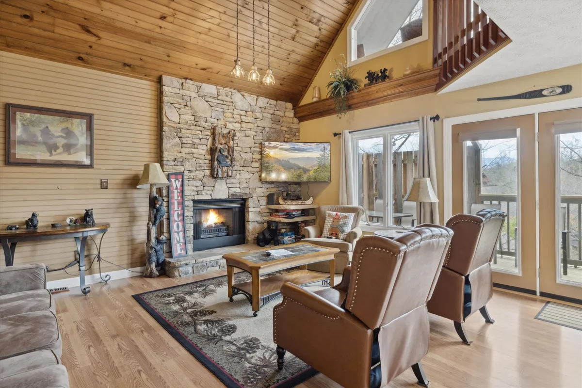 A welcoming living area featuring a stone fireplace, large windows that frame the Smoky Mountains, a wooden ceiling, and plush, comfortable seating arranged for group gatherings. The atmosphere is cozy and inviting, perfect for laughter and conversation by the fire with scenic views just outside.