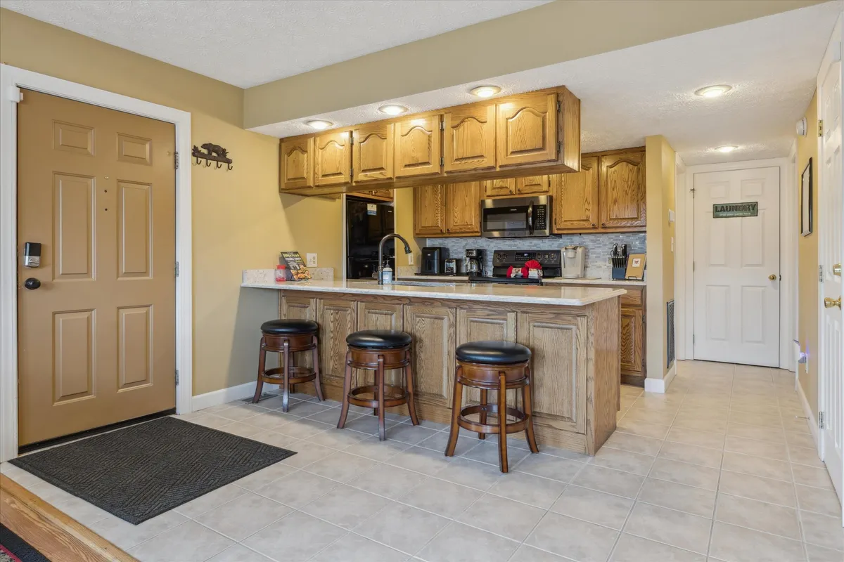 A well-equipped kitchen with a central island, wooden cabinets, bar stools for casual seating, and a welcoming front door nearby. The space is designed for both cooking and entertaining, with plenty of room for group meal prep and relaxed breakfasts.